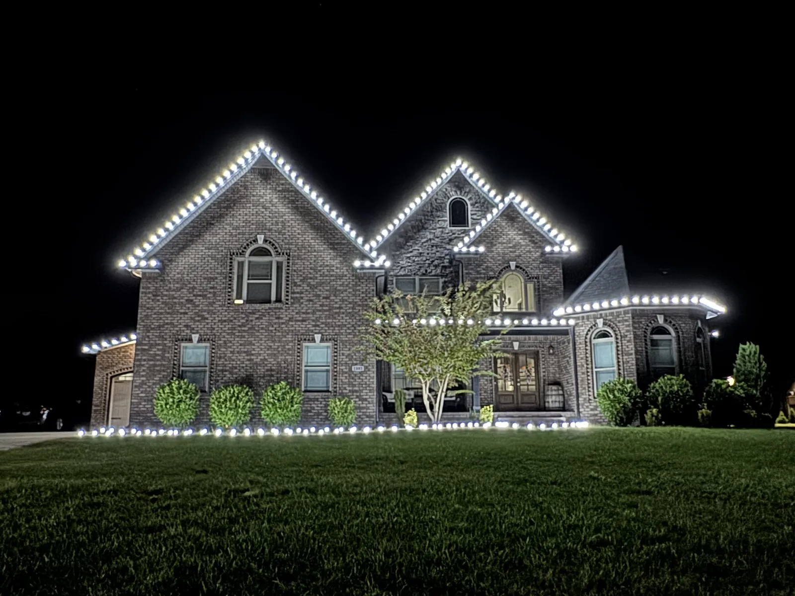 Cool white lights outlining roof peaks and shrubs of a brick home